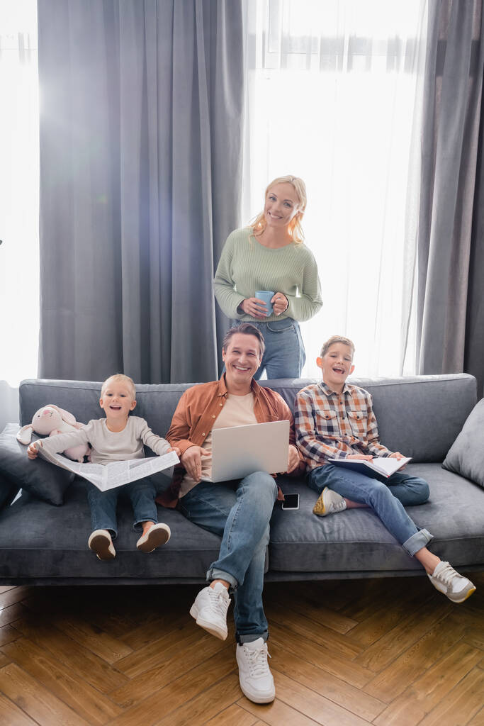 glückliche Frau mit Tee, Mann mit Laptop und Jungen mit Zeitung und Notizbuch, die im Wohnzimmer in die Kamera schauen - Foto, Bild
