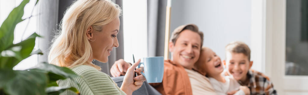 mujer feliz beber té cerca de la familia borrosa en la sala de estar, pancarta - Foto, imagen