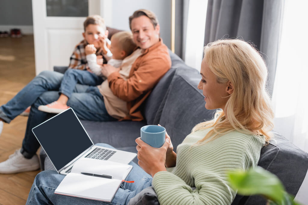 mulher com notebook, laptop e xícara de chá sentado no sofá perto de família alegre borrada - Foto, Imagem