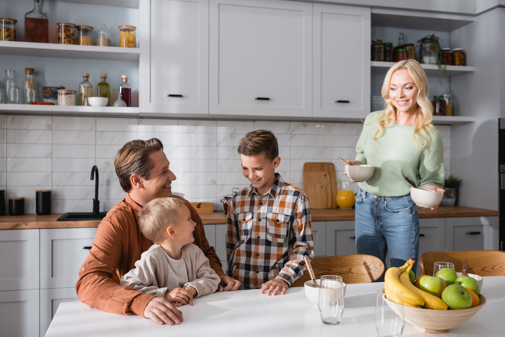 femme souriante tenant des bols près du mari et des fils assis à la table de cuisine - Photo, image