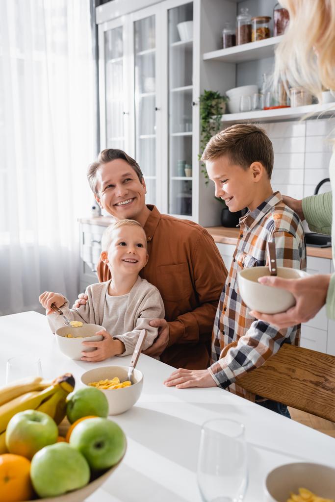 alegre niño comiendo copos de maíz cerca de padres y hermano en la cocina - Foto, imagen