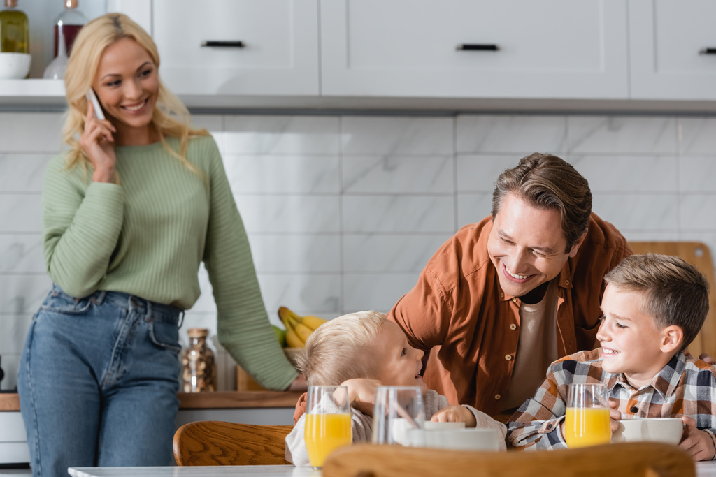 verschwommene Frau telefoniert neben glücklichem Ehemann, der neben Söhnen steht und in Küche frühstückt - Foto, Bild