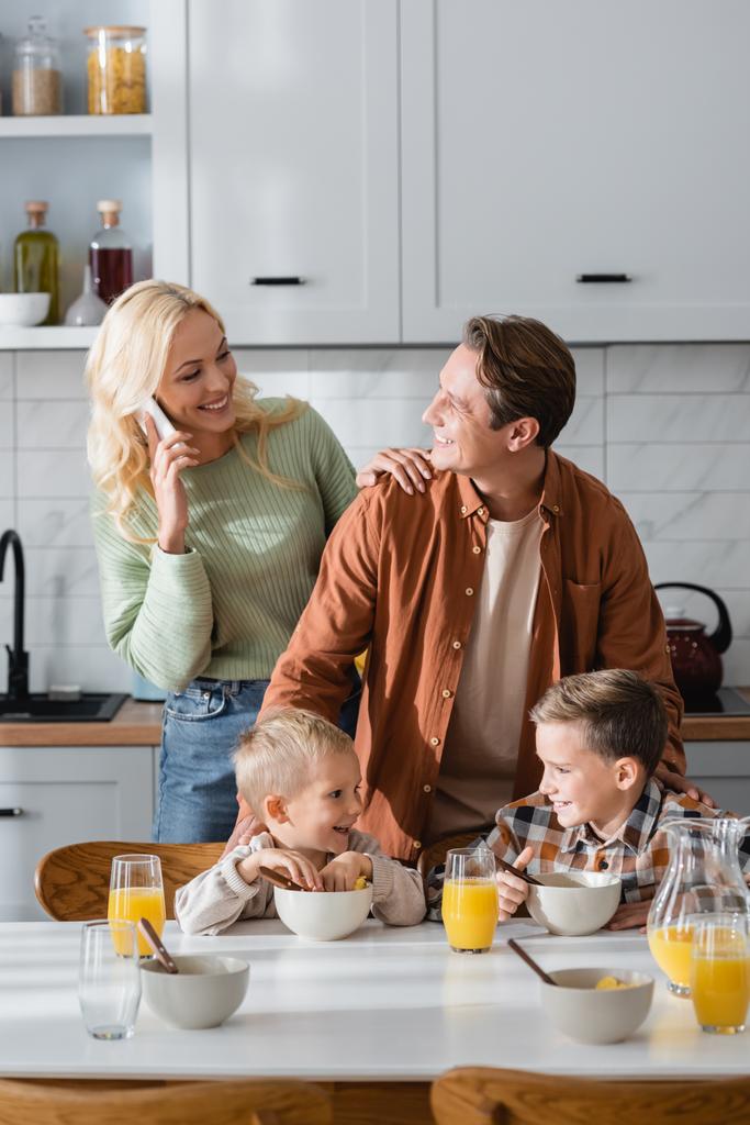 mulher sorrindo falando no telefone celular enquanto olha para o marido e os meninos durante o café da manhã - Foto, Imagem