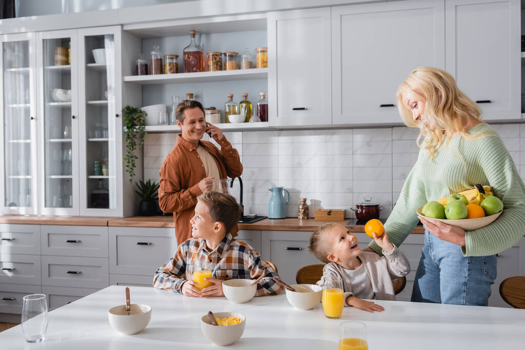 smiling man talking on smartphone while wife holding fruits near kids in kitchen - Photo, Image