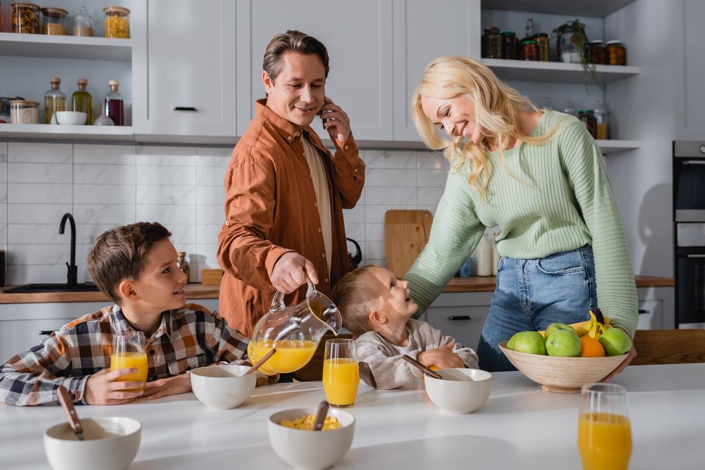 man talking on smartphone while pouring orange juice during breakfast with family - Photo, Image
