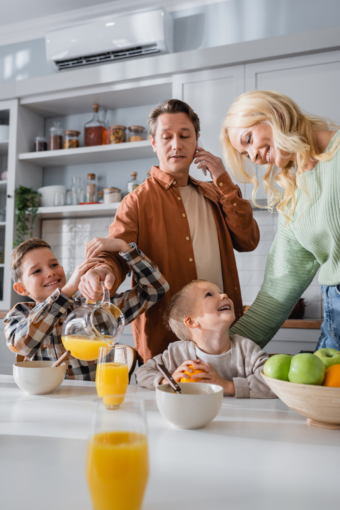 muž nalévání pomerančový džus při rozhovoru na smartphone během snídaně s rodinou - Fotografie, Obrázek