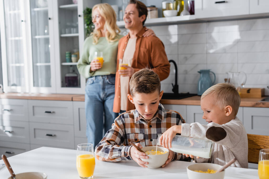 menino derramando leite em flocos de milho enquanto café da manhã com o irmão perto da família turva - Foto, Imagem