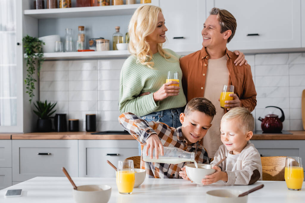 couple joyeux avec des verres de jus d'orange près des enfants qui prennent le petit déjeuner dans la cuisine - Photo, image