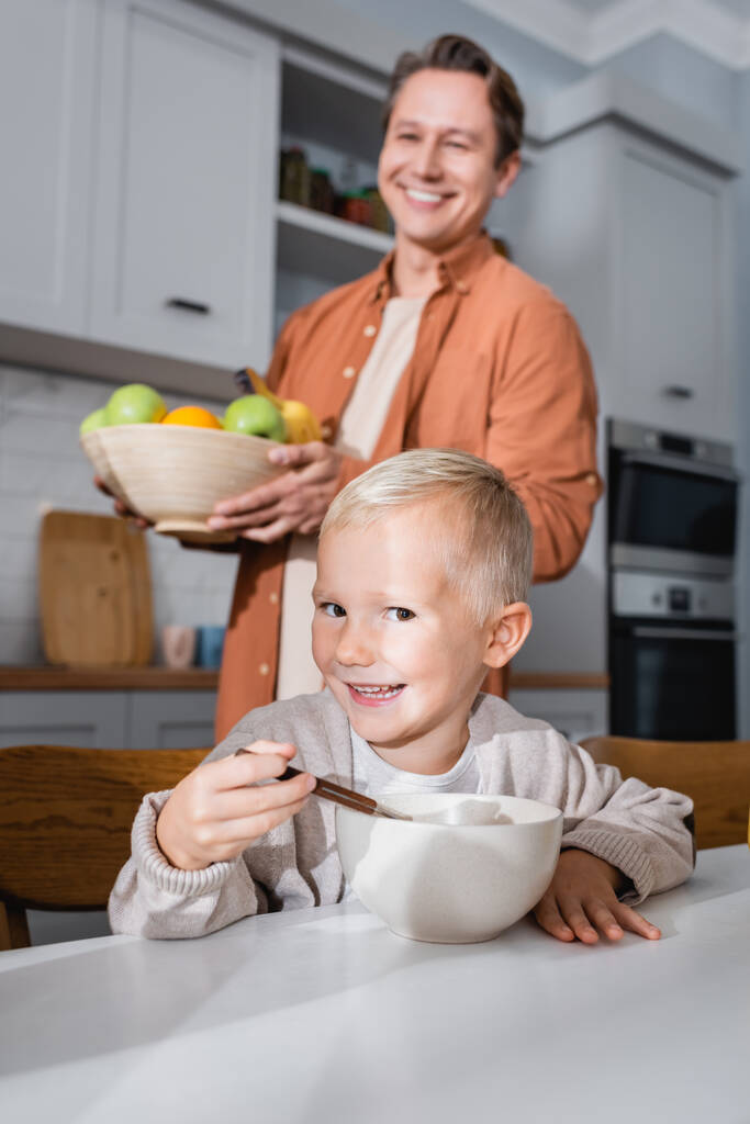 menino sorrindo olhando para a câmera enquanto toma café da manhã perto de pai desfocado com frutas frescas - Foto, Imagem