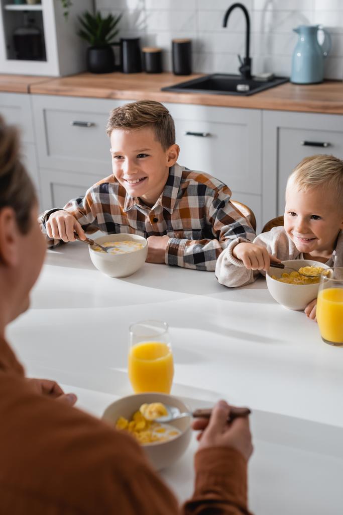iloinen pojat syövät maukkaita maissihiutaleita lähellä hämärtynyt isä keittiössä - Valokuva, kuva
