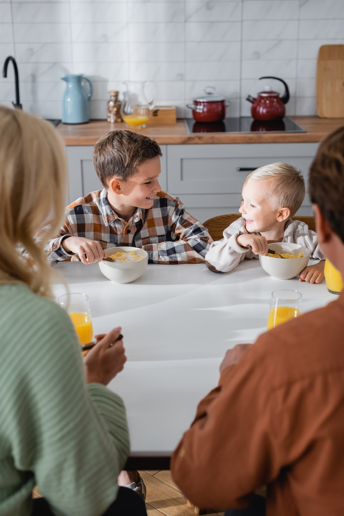 usmívající se kluci mají snídani při jídle kukuřičné vločky v blízkosti rozmazaných rodičů - Fotografie, Obrázek
