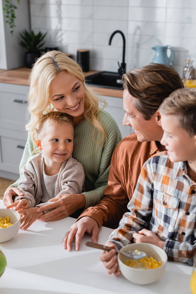 famille heureuse regardant l'autre près des bols avec des flocons de maïs sur la table de cuisine - Photo, image