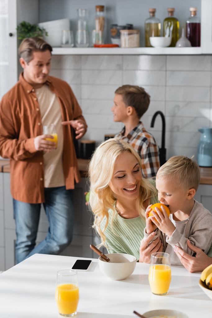 niño complacido comiendo naranja cerca de mamá y papá borroso con el hermano en la cocina - Foto, imagen
