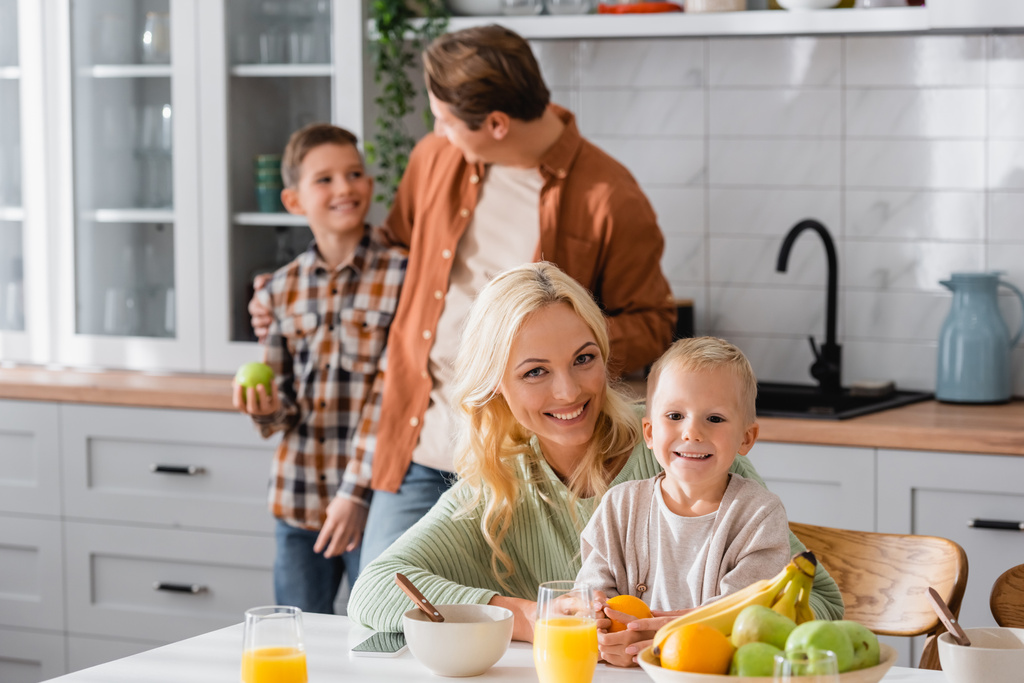 šťastná žena při pohledu na kameru při snídani s dítětem v blízkosti rozmazaný manžel a syn - Fotografie, Obrázek
