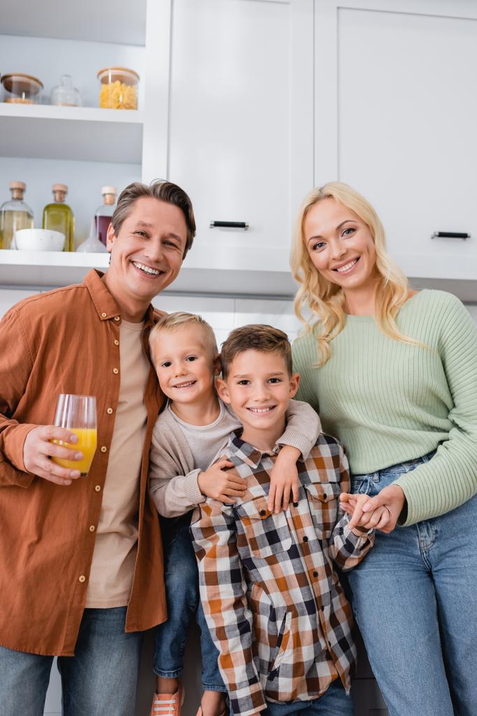 happy man with orange juice smiling at camera near wife and sons embracing in kitchen - Photo, Image