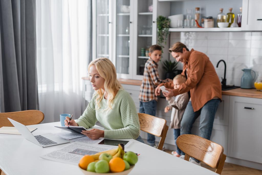 busy woman writing in notebook while working near laptop and blurred family in kitchen - Photo, Image