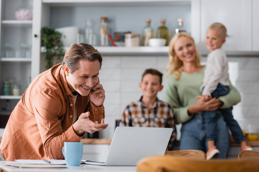férfi beszél okostelefonon, miközben dolgozik közelében laptop és elmosódott család a konyhában - Fotó, kép