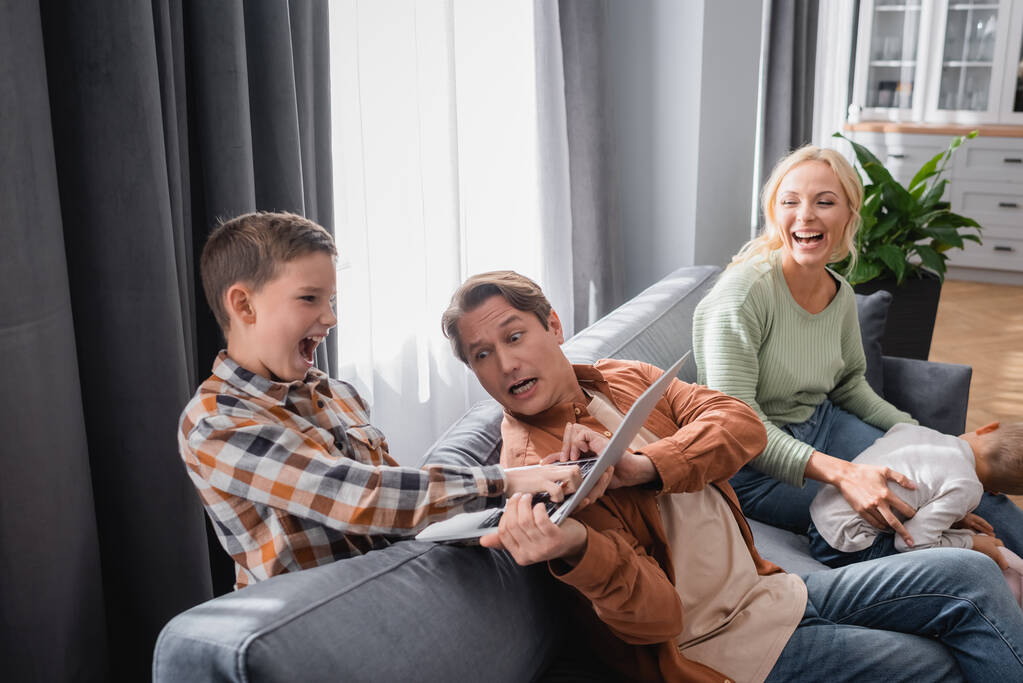 aufgeregter Junge nimmt Laptop vom geschäftigen Vater neben Mutter lachend auf Couch - Foto, Bild