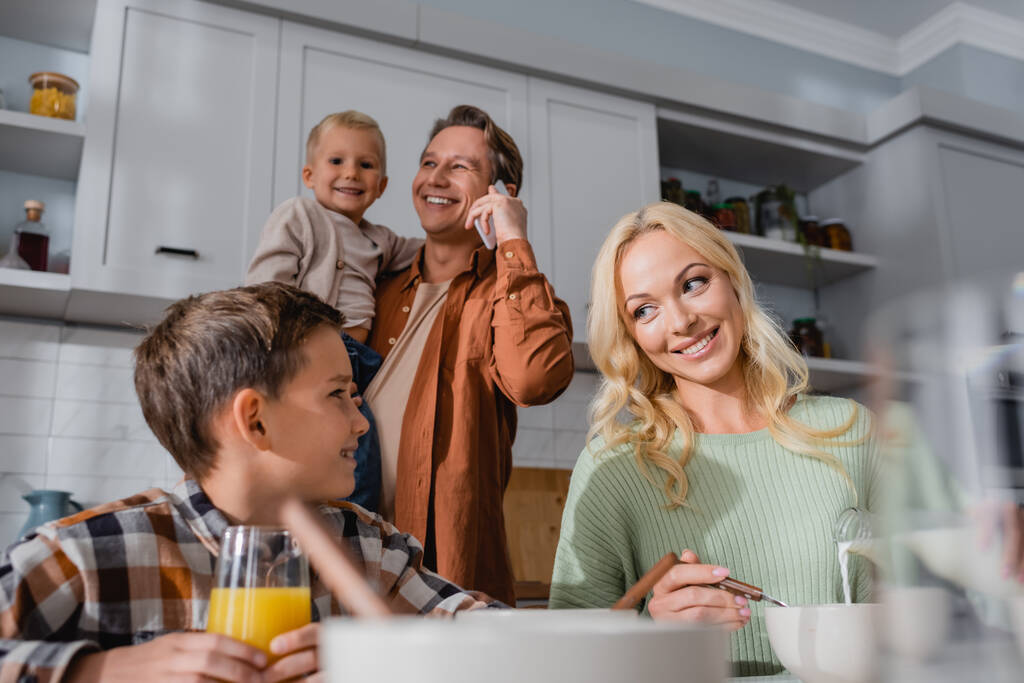 smiling man holding kid and talking on smartphone near wife and son having breakfast in kitchen - Photo, Image