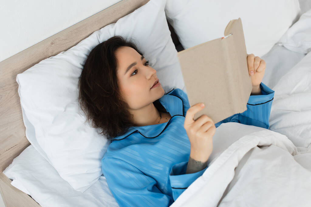 προβολή υψηλής γωνίας του βιβλίου μελαχρινή γυναίκα ανάγνωση στο κρεβάτι - Φωτογραφία, εικόνα
