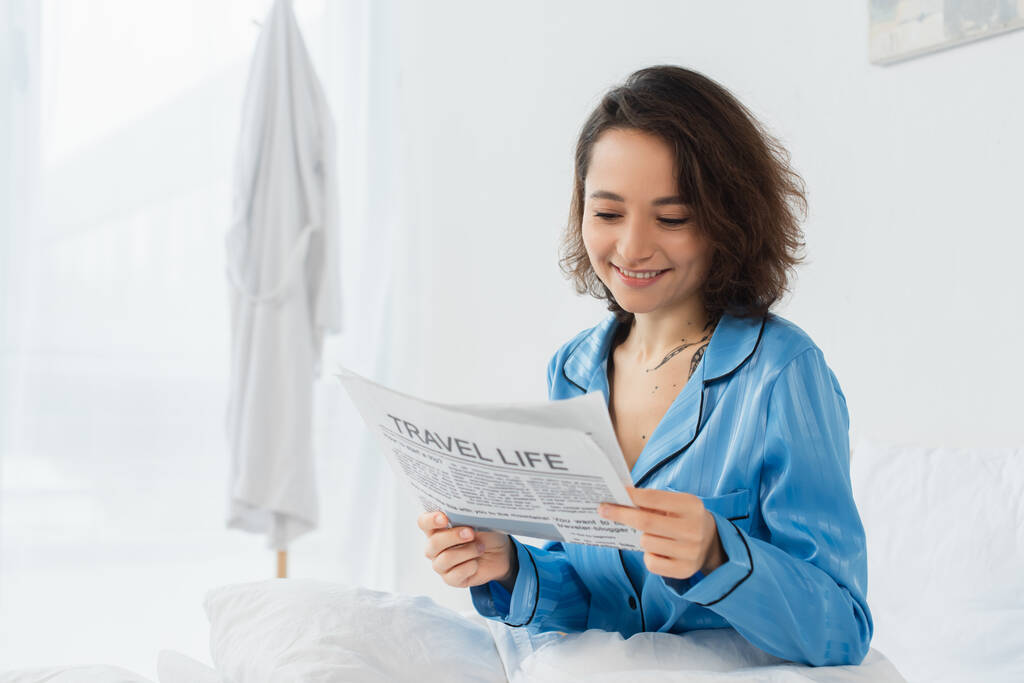 mujer feliz en pijama azul leyendo la vida de viaje periódico en la cama - Foto, imagen