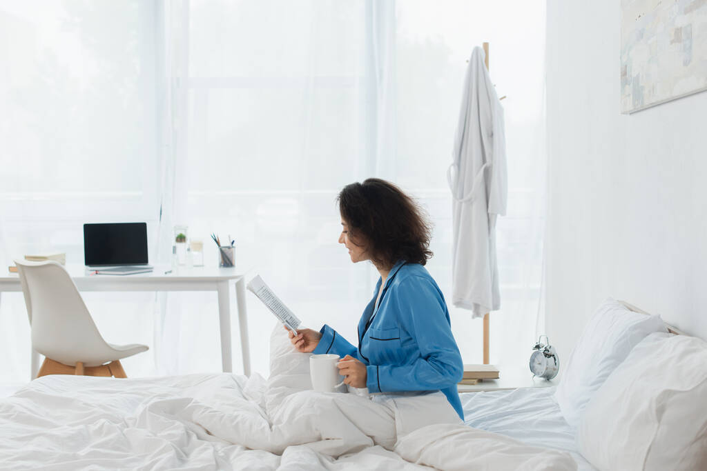 新聞を読んだりベッドでカップを持っている青いパジャマ姿の陽気な女性 - 写真・画像