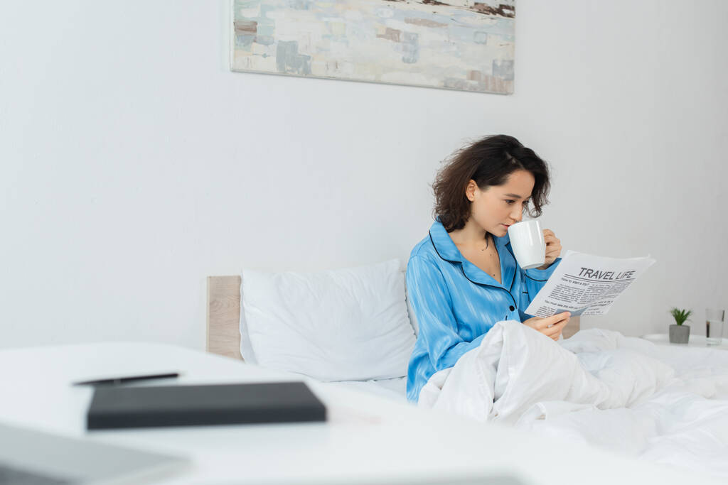 brunetka kobieta w niebieski piżama czytanie podróże życie gazeta i picie herbaty w łóżku - Zdjęcie, obraz