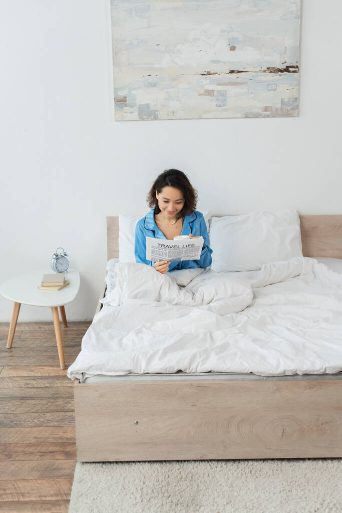веселая молодая женщина в пижаме читает газету о путешествиях и держит кубок в постели - Фото, изображение