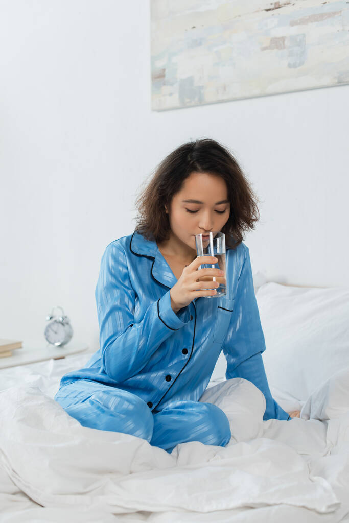 jovem mulher de pijama azul segurando vidro de água no quarto  - Foto, Imagem