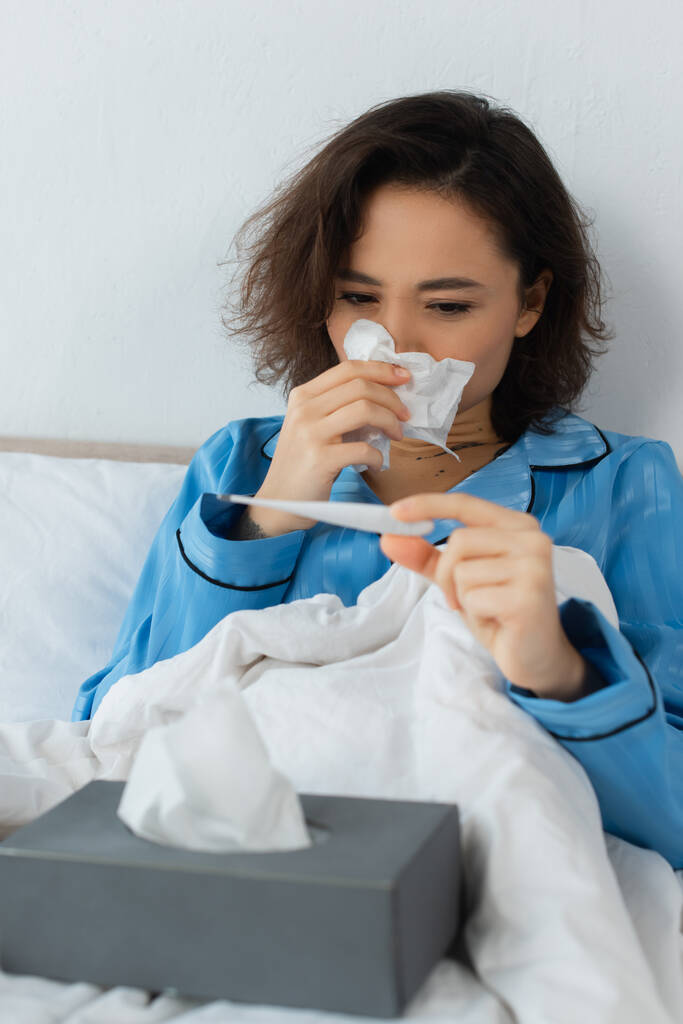 больная молодая женщина чихает в ткани и смотрит на электронный термометр - Фото, изображение