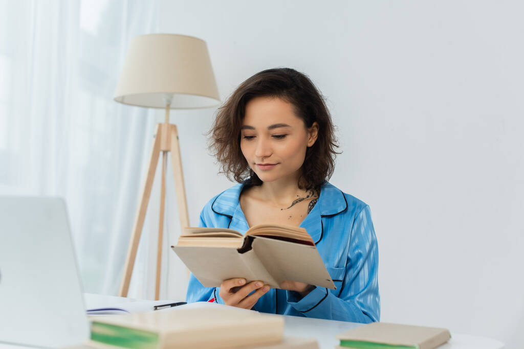 tatoué jeune femme lecture livre près ordinateur portable sur le bureau - Photo, image