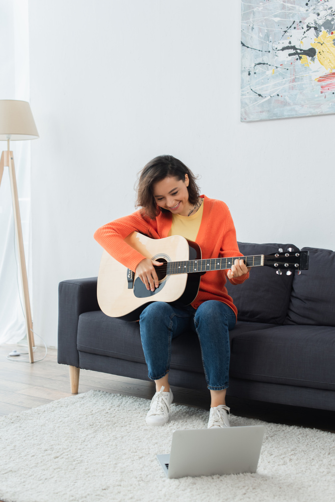 fröhliche junge Frau lernt Akustikgitarre neben Laptop auf Teppich zu spielen - Foto, Bild