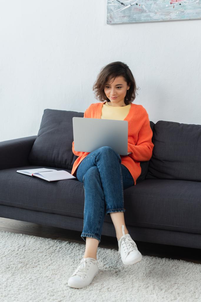 брюнетка молодая женщина с ноутбуком рядом с ноутбуком на диване - Фото, изображение