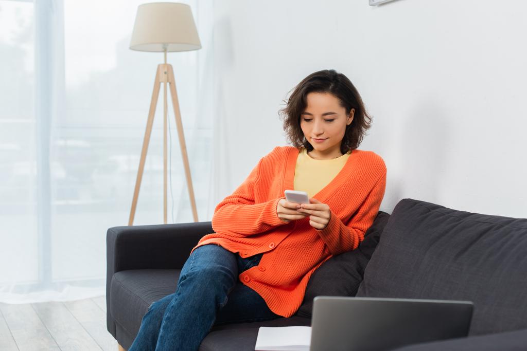 mujer joven mensajes de texto en el teléfono celular cerca de la computadora portátil en la sala de estar  - Foto, imagen