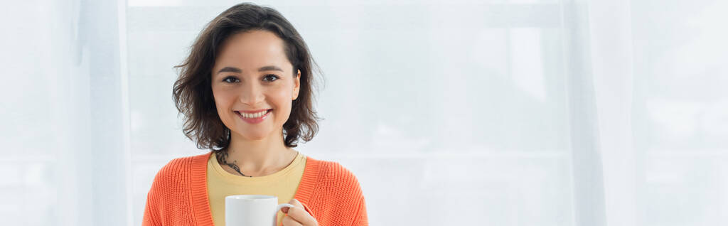 pleased young woman smiling and holding cup near white curtain, banner - Photo, Image