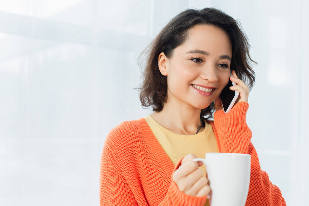pozitív fiatal nő gazdaság csésze és beszél okostelefon közelében fehér függöny  - Fotó, kép