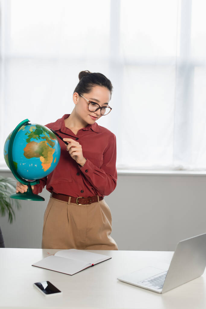 young cheerful teacher in eyeglasses holding globe and looking at laptop near smartphone on desk - Photo, Image