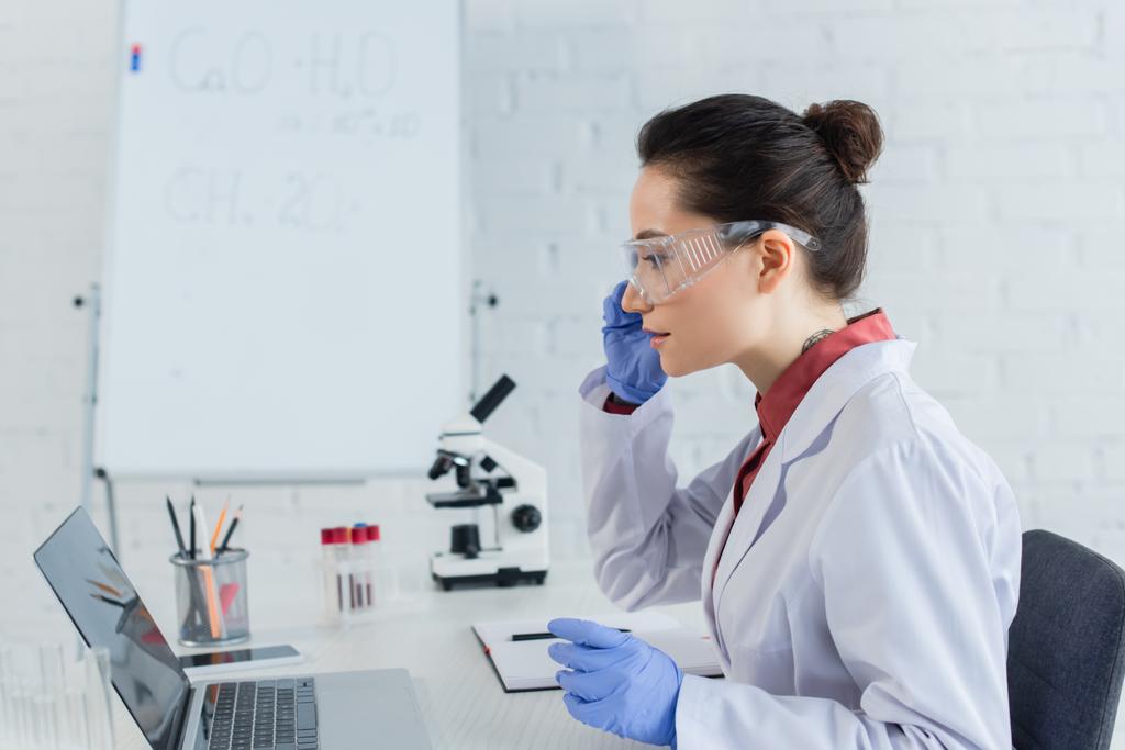 zijaanzicht van getatoeëerde wetenschapper in witte jas en bril kijkend naar laptop in de buurt van reageerbuizen  - Foto, afbeelding