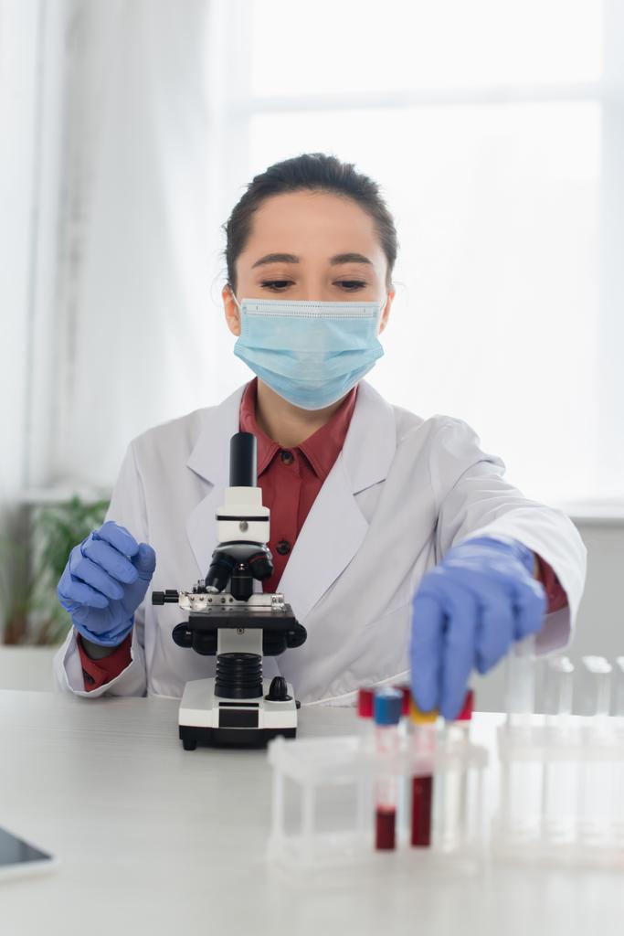 jeune scientifique en gants de latex et masque médical atteignant les éprouvettes près du microscope flou - Photo, image