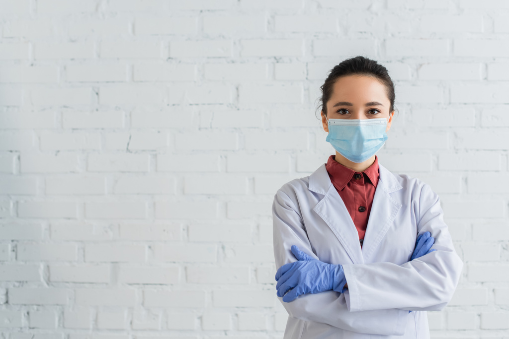 брюнетка лікар в медичній масці дивиться на камеру і стоїть з схрещеними руками
 - Фото, зображення