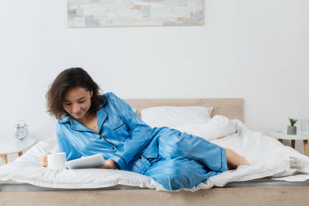 mujer complacida en pijama azul sosteniendo una taza de café y usando una tableta digital en el dormitorio  - Foto, Imagen