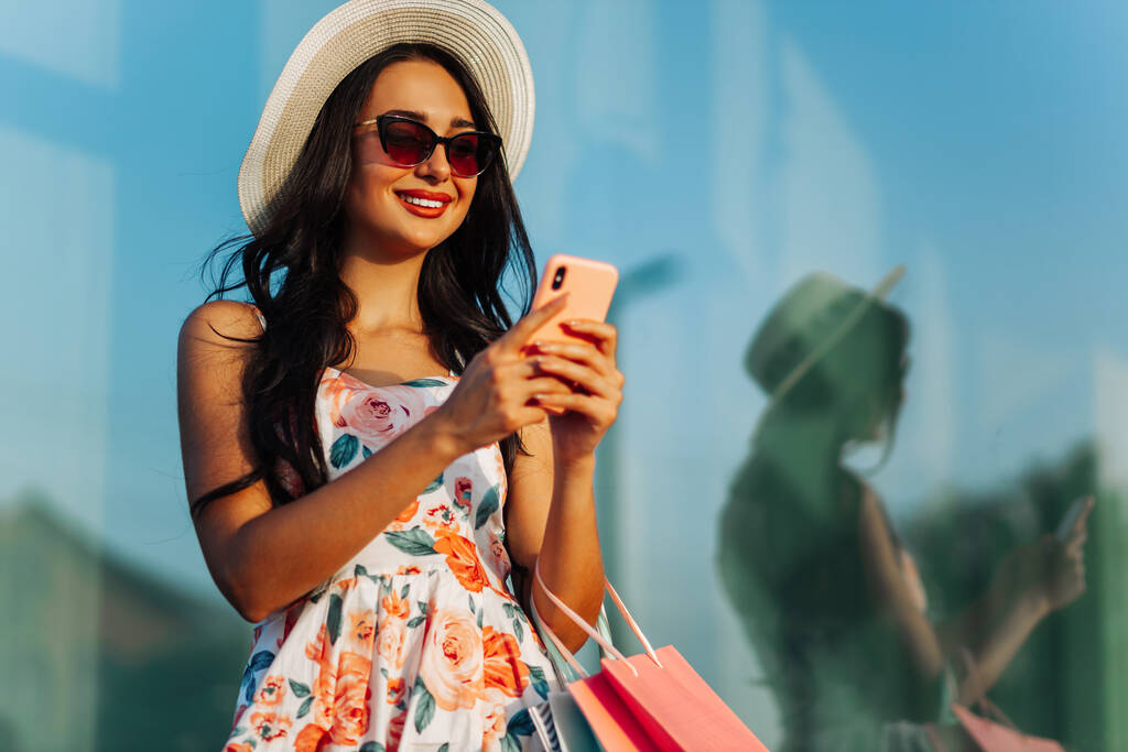 modieuze jonge vrouw, in een mooie jurk en zonnebril, tassen met aankopen, met een mobiele telefoon en glimlach, winkelen in een damesboetiek op Black Friday, Big Sale, Winkelen - Foto, afbeelding
