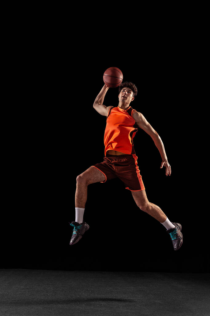 Retrato completo de entrenamiento de jugador de baloncesto aislado en el fondo oscuro del estudio. Alto atleta muscular saltando con la pelota. - Foto, imagen