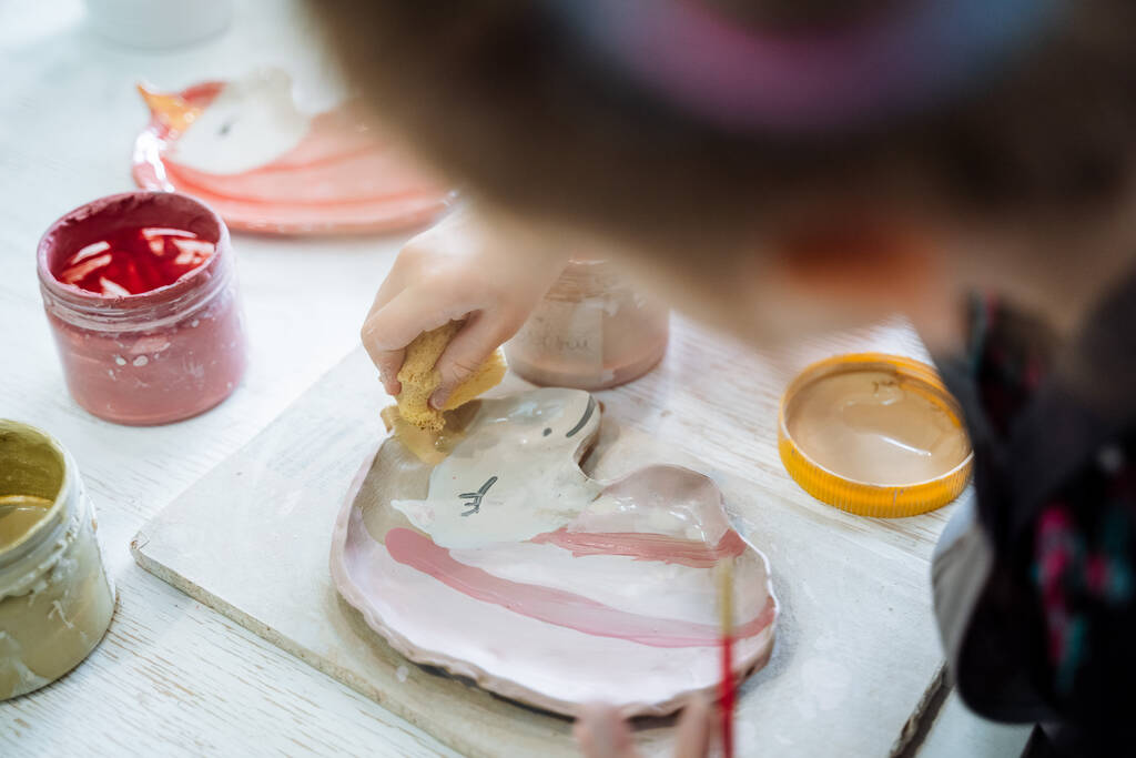 Tyttö maalaa lautasellisen savea vaaleanpunaisen yksisarvisen muotoiseksi. Luovuus keramiikka työpajassa. Laadukas kuva - Valokuva, kuva