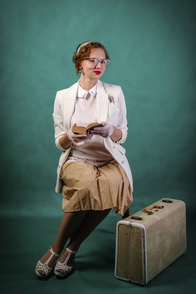 Jovem com óculos, vestida em estilo retrô, sentada em uma cadeira, com uma mala aos pés, com um livro nas mãos. Retrato de estúdio - Foto, Imagem