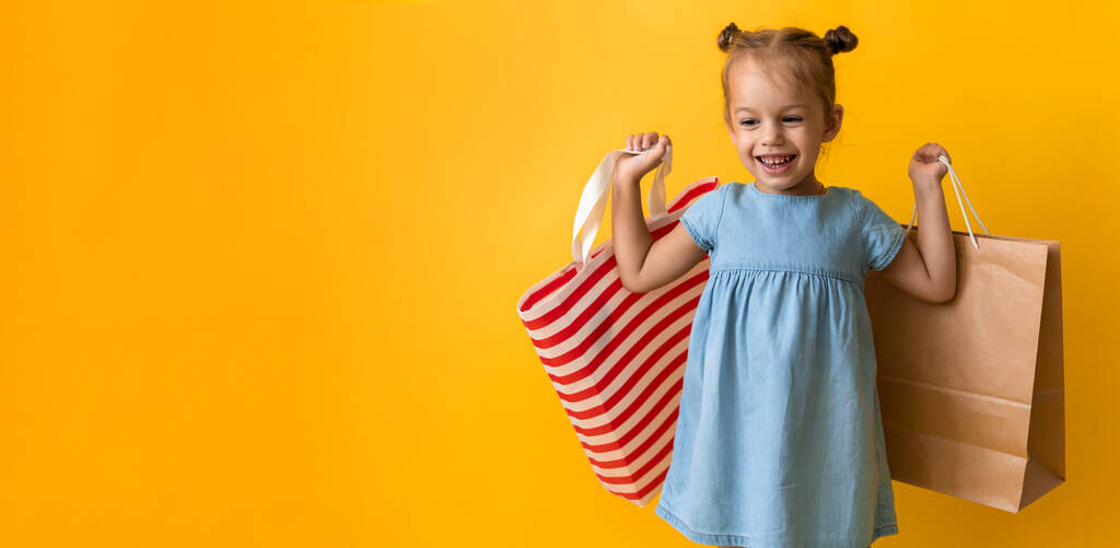 Баннерный портрет Кавказка Красивая счастливая маленькая дошкольница улыбается весело и держа картонные сумки, изолированные на оранжевом желтом фоне. Счастье, Потребительство, Продажа людей торговый концепт - Фото, изображение