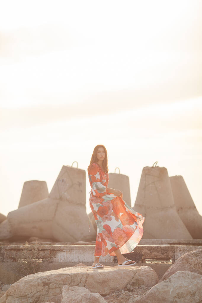Красивая женщина в длинном красном платье рядом с волнорезом, прогулка у моря - Фото, изображение