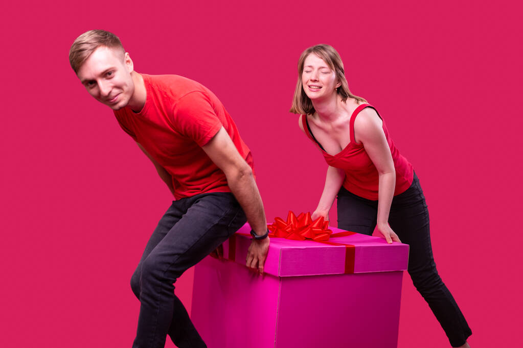 Mladý usmívající se muž a žena s obličejovým výrazem v červených tričkách a černých džínách táhnoucí krabici růžové barvy a mašle. - Fotografie, Obrázek