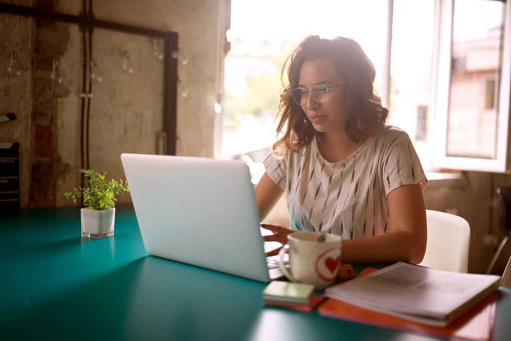 Egy fiatal nő dolgozik egy laptopon, miközben az íróasztalnál ül kellemes légkörben az irodában. - Fotó, kép