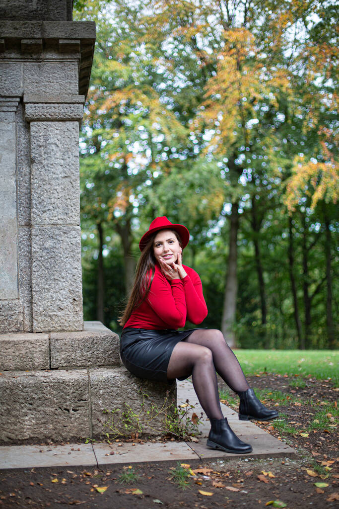 Сидящая белая женщина с длинными темными волосами в ярко-красном у памятника в парке - Фото, изображение